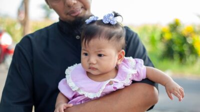Seorang ayah tersenyum bahagia sambil menggendong anak perempuannya dengan penuh kasih sayang.