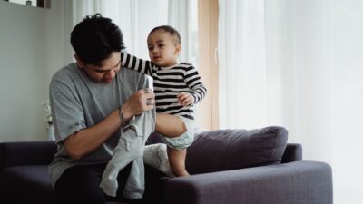 A father helping his toddler get dressed on a couch, illustrating a moment for positive perubahan gaya parenting.