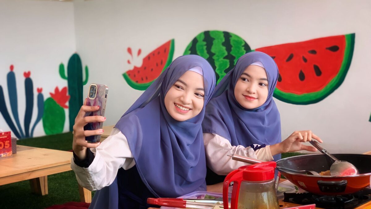Two young women in blue hijabs smiling while taking a selfie together indoors.