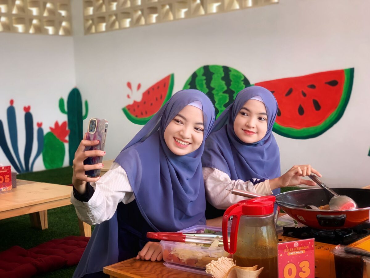 Two young women in blue hijabs smiling while taking a selfie together indoors.