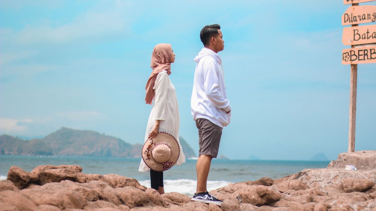 A young couple stands on a rocky beach overlooking the sea beside a signpost.