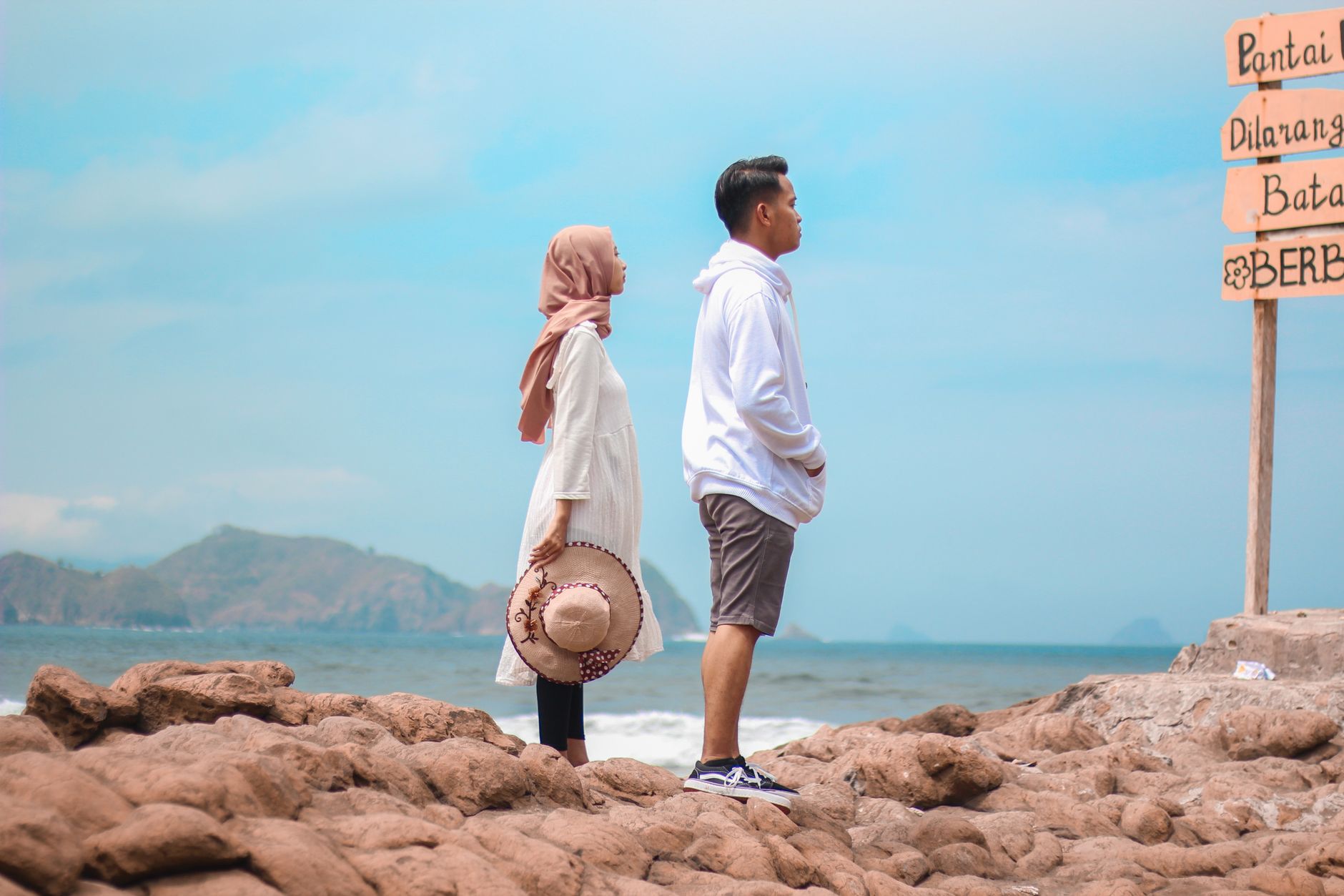 A young couple stands on a rocky beach overlooking the sea beside a signpost.