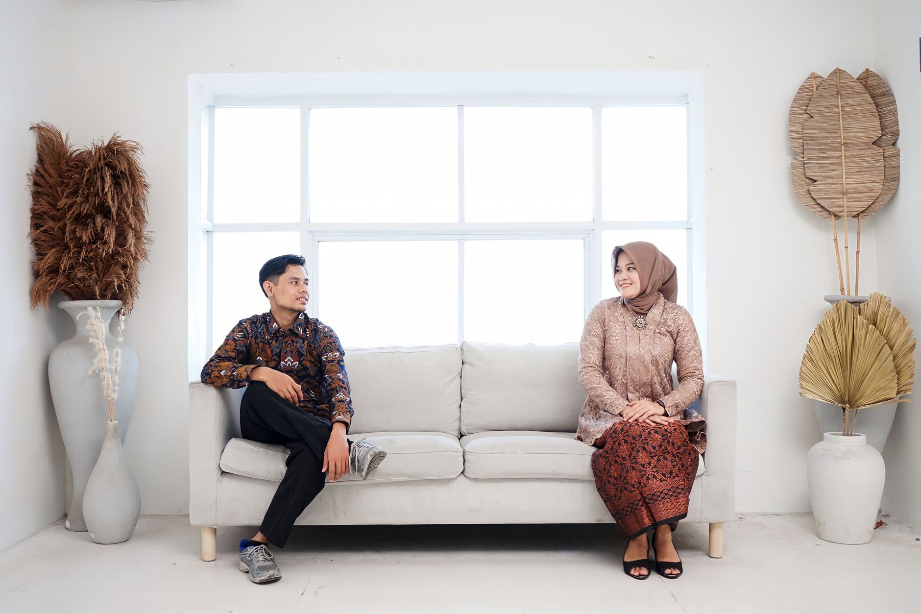 Pasangan extrovert introvert mengenakan pakaian adat Indonesia, duduk terpisah di sofa abu-abu dalam ruangan terang.