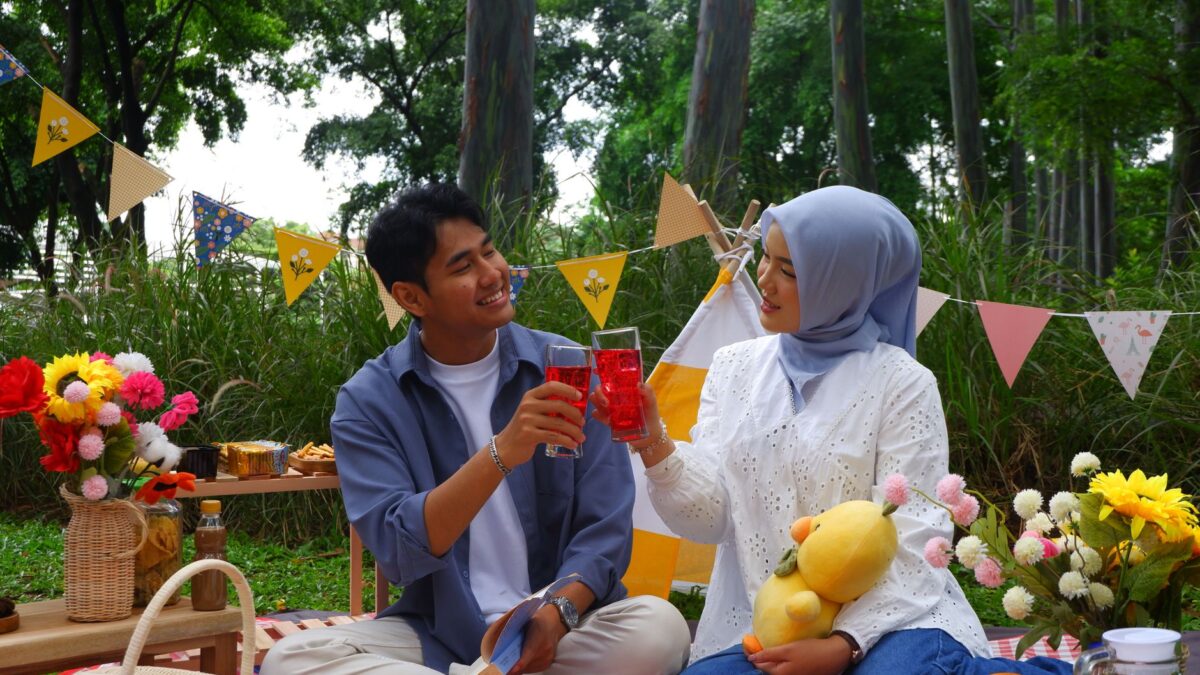 A young Muslim couple clinks glasses during a picnic in a park while memulai kencan kembali.
