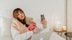 Tired young woman in bed holding a smartphone, illustrating the Fenomena FOMO.