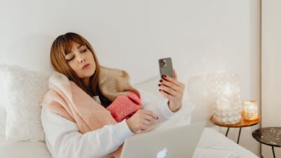Tired young woman in bed holding a smartphone, illustrating the Fenomena FOMO.