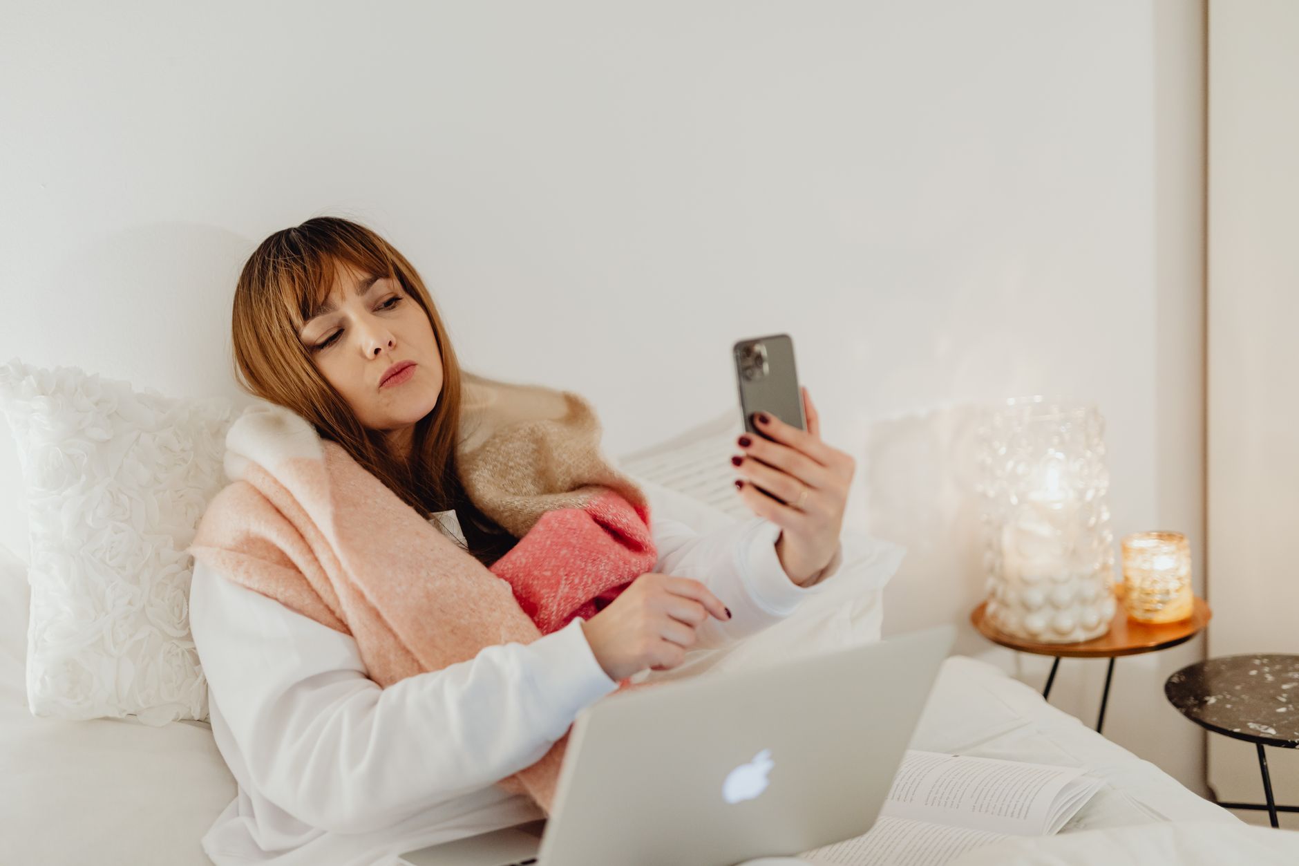 Tired young woman in bed holding a smartphone, illustrating the Fenomena FOMO.