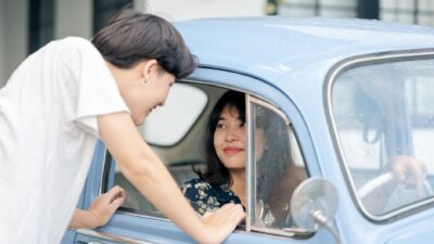 A young woman in a white t-shirt talks to a woman sitting inside a classic light blue car.