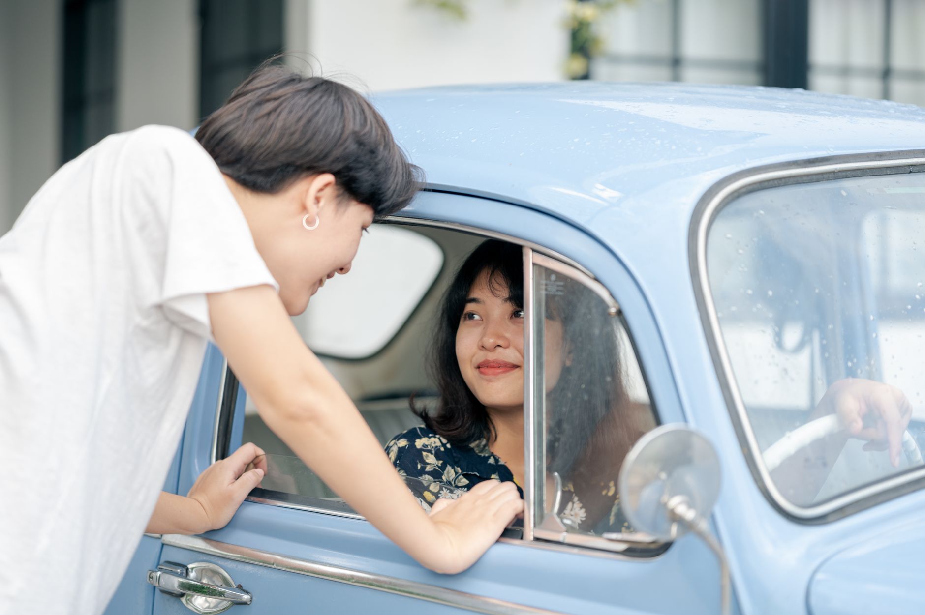 A young woman in a white t-shirt talks to a woman sitting inside a classic light blue car.