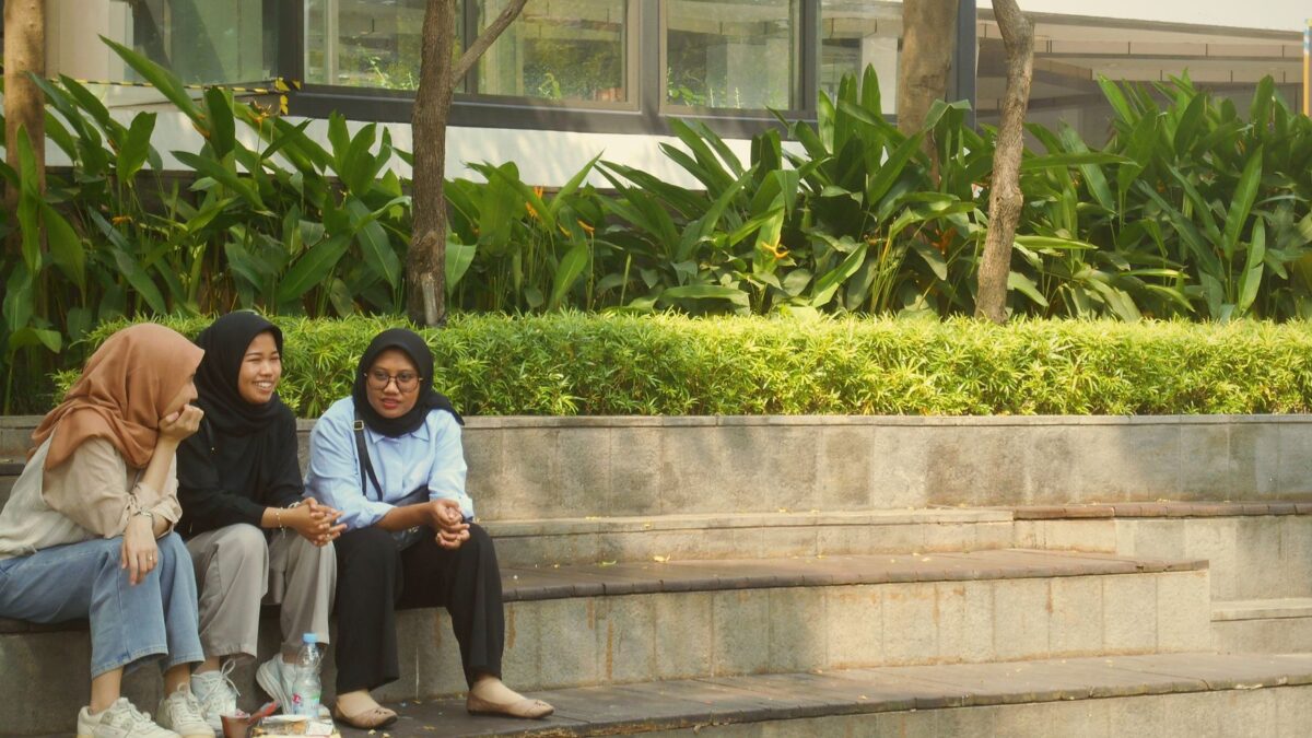 Tiga wanita berhijab duduk di tangga luar ruangan sambil mengobrol, terkait persepsi waktu santai.