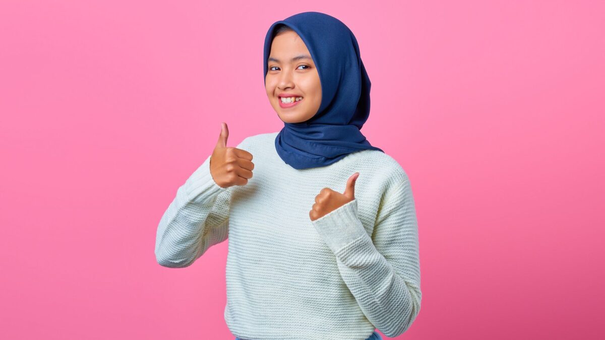 A smiling woman in a blue hijab and white sweater gives a thumbs-up against a pink background.