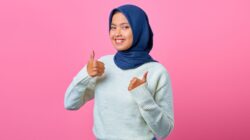 A smiling woman in a blue hijab and white sweater gives a thumbs-up against a pink background.
