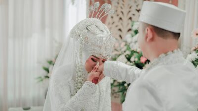 Pengantin wanita Indonesia berhijab putih mencium tangan suami dengan busana pernikahan tradisional.