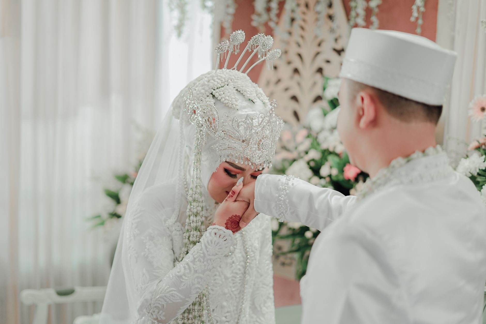 Pengantin wanita Indonesia berhijab putih mencium tangan suami dengan busana pernikahan tradisional.