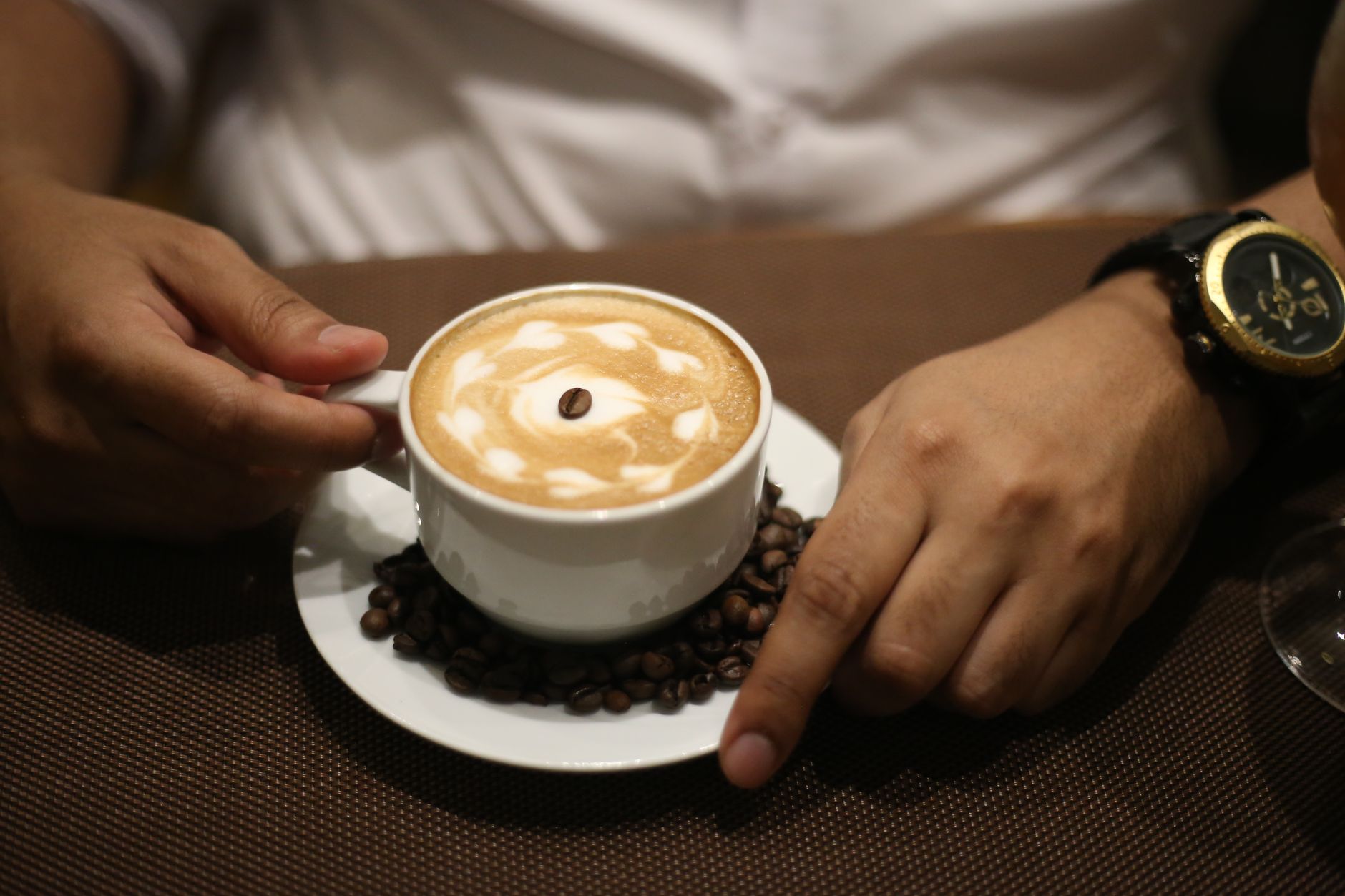 Seseorang memegang secangkir cappuccino dengan latte art dan biji kopi di sekitarnya sebagai ilustrasi bahaya konsumsi kafein