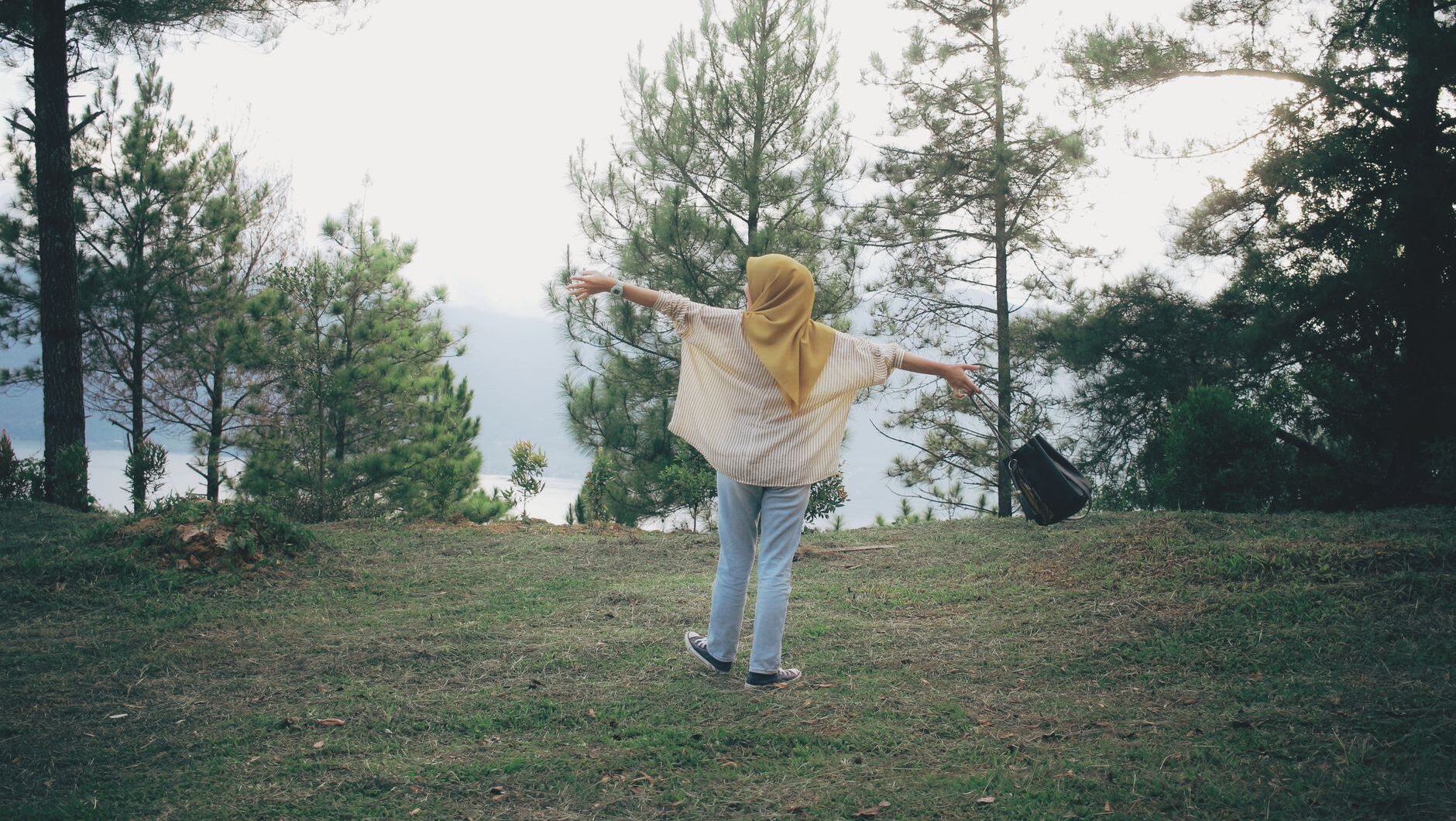 A woman in a hijab with arms outstretched, enjoying scenic forest views for hidup bahagia.