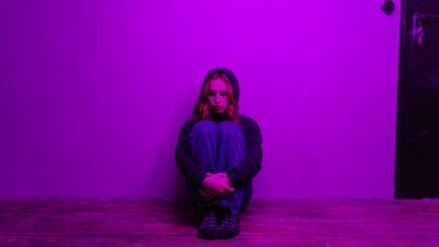 A lonely young woman sits hugging her knees against a wall in a dimly lit room, representing depression.