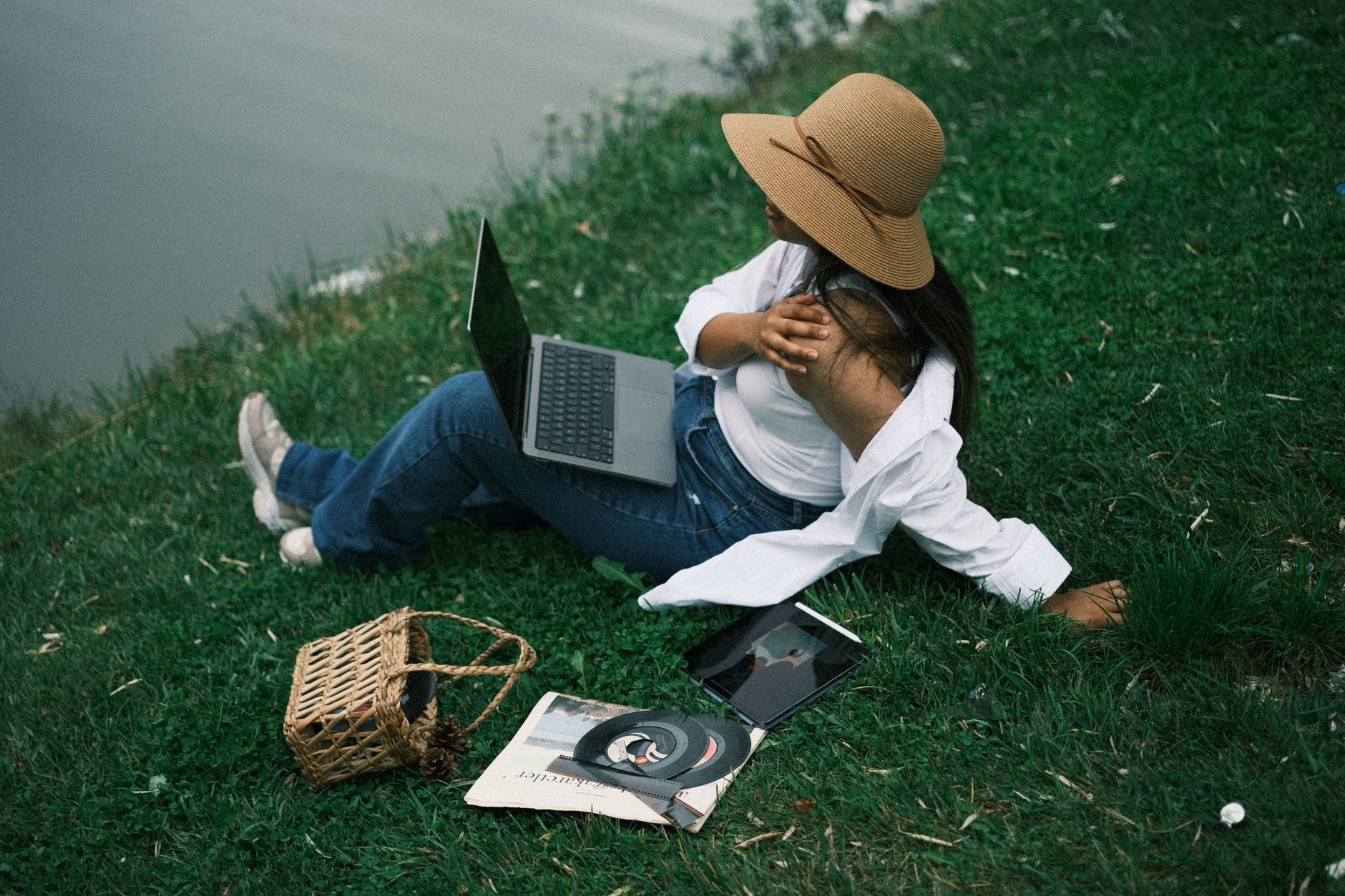 A woman in a straw hat sits on green grass by the water, using a laptop for a digital detox.