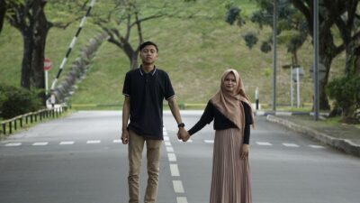 Seasang kekasih berjalan sambil bergandengan tangan di jalan berpaving yang dikelilingi pepohonan sebagai bukti cinta sejati.