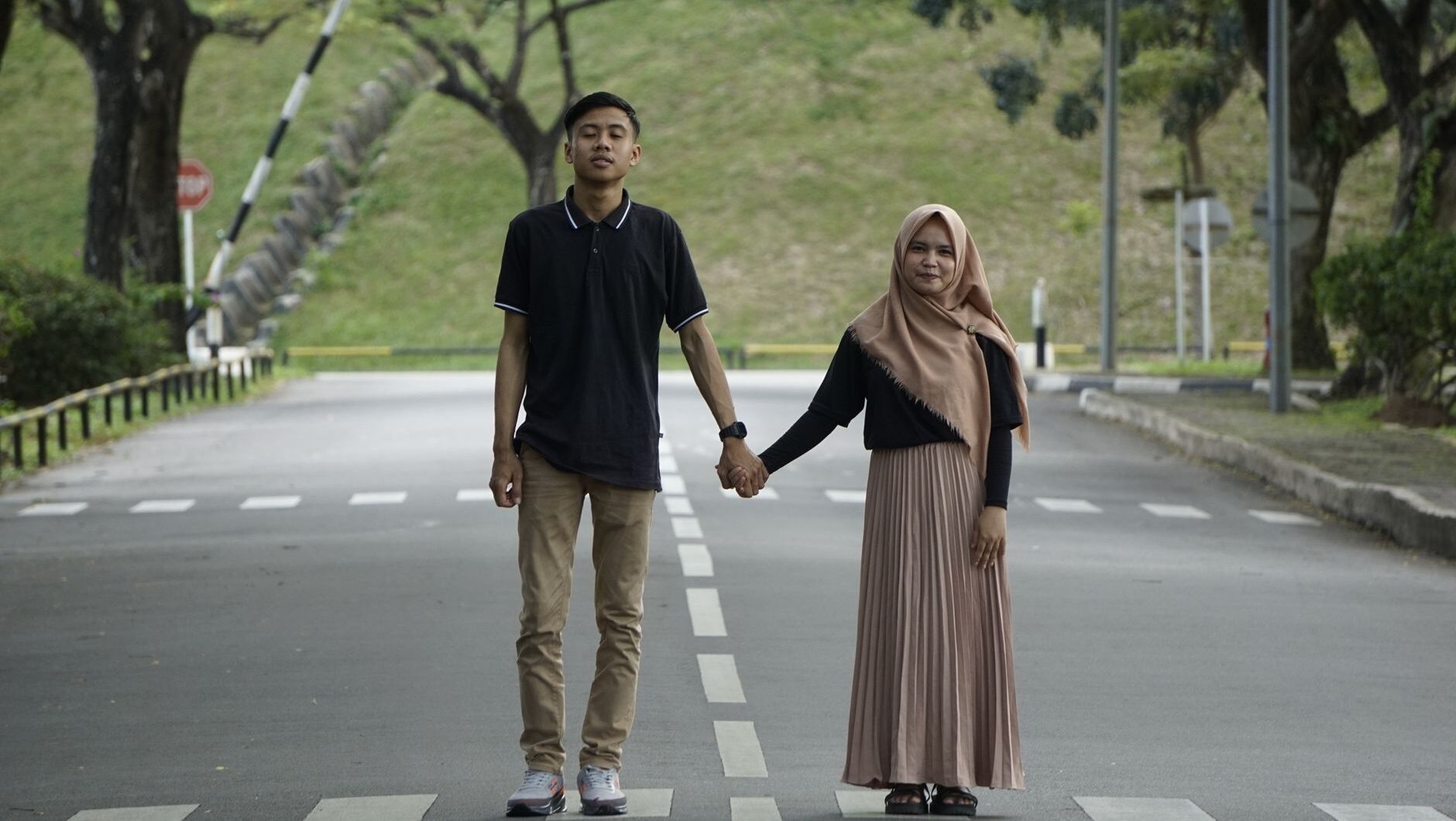 A young couple holds hands while walking together on a paved road surrounded by lush green trees, representing a true pasanga