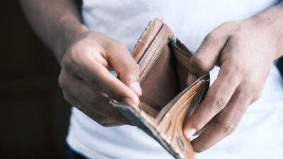 Tangan seseorang membuka dompet kulit cokelat yang sudah usang dan tampak kosong tanpa uang tunai.