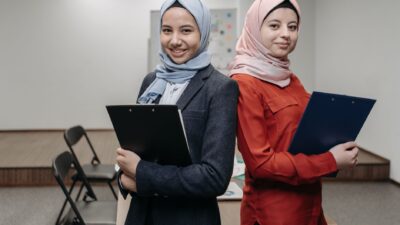 Two smiling young women in hijabs pose back-to-back in an office holding clipboards, illustrating good etika kerja.