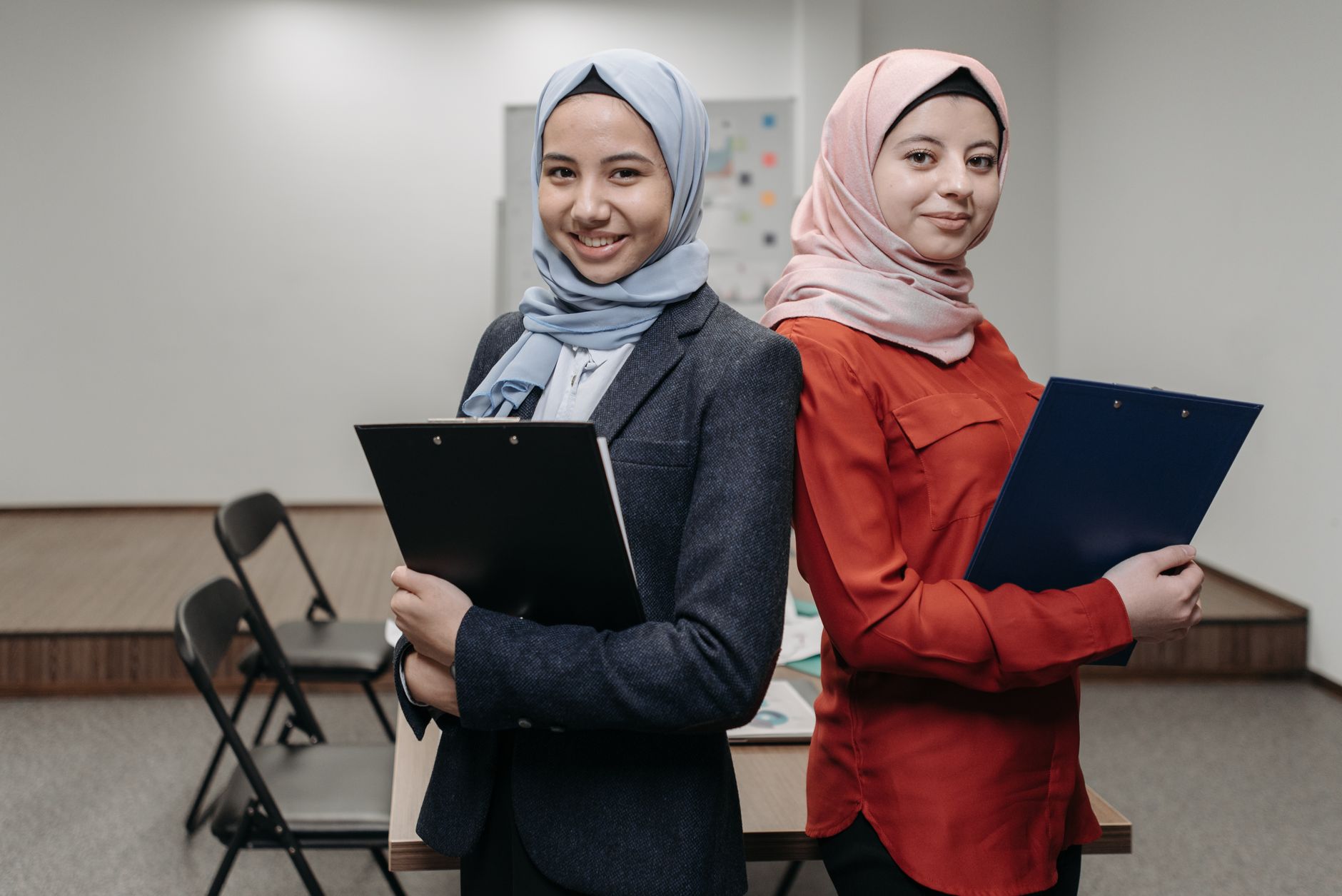 Two smiling young women in hijabs pose back-to-back in an office holding clipboards, illustrating good etika kerja.