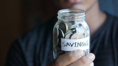 A person holding a glass savings jar filled with coins, representing tips menabung efektif.