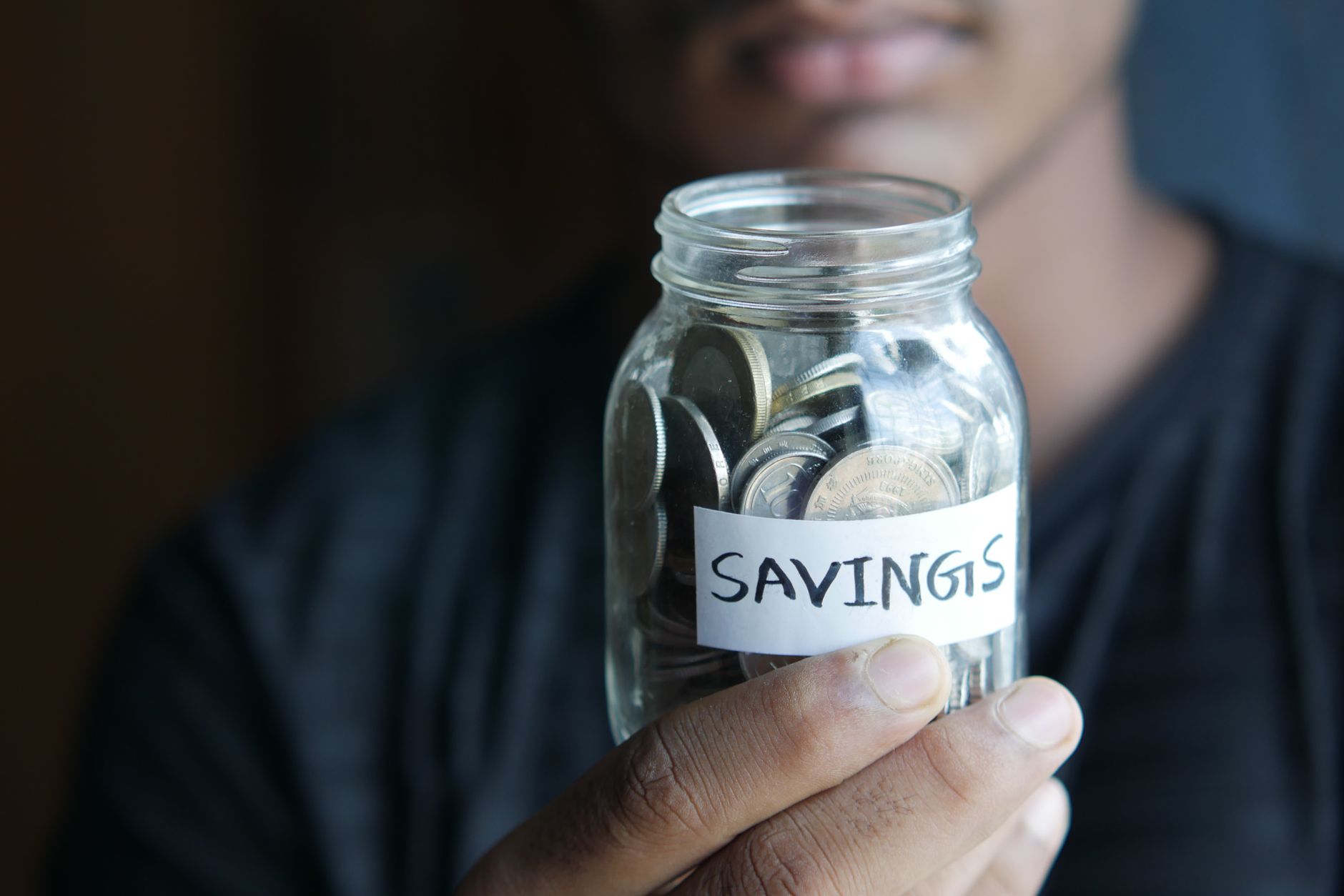 A person holding a glass savings jar filled with coins, representing tips menabung efektif.