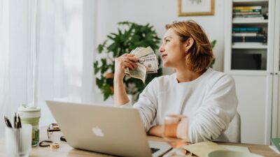 A woman at a desk with a laptop holding cash and looking thoughtfully away for a side hustle concept.