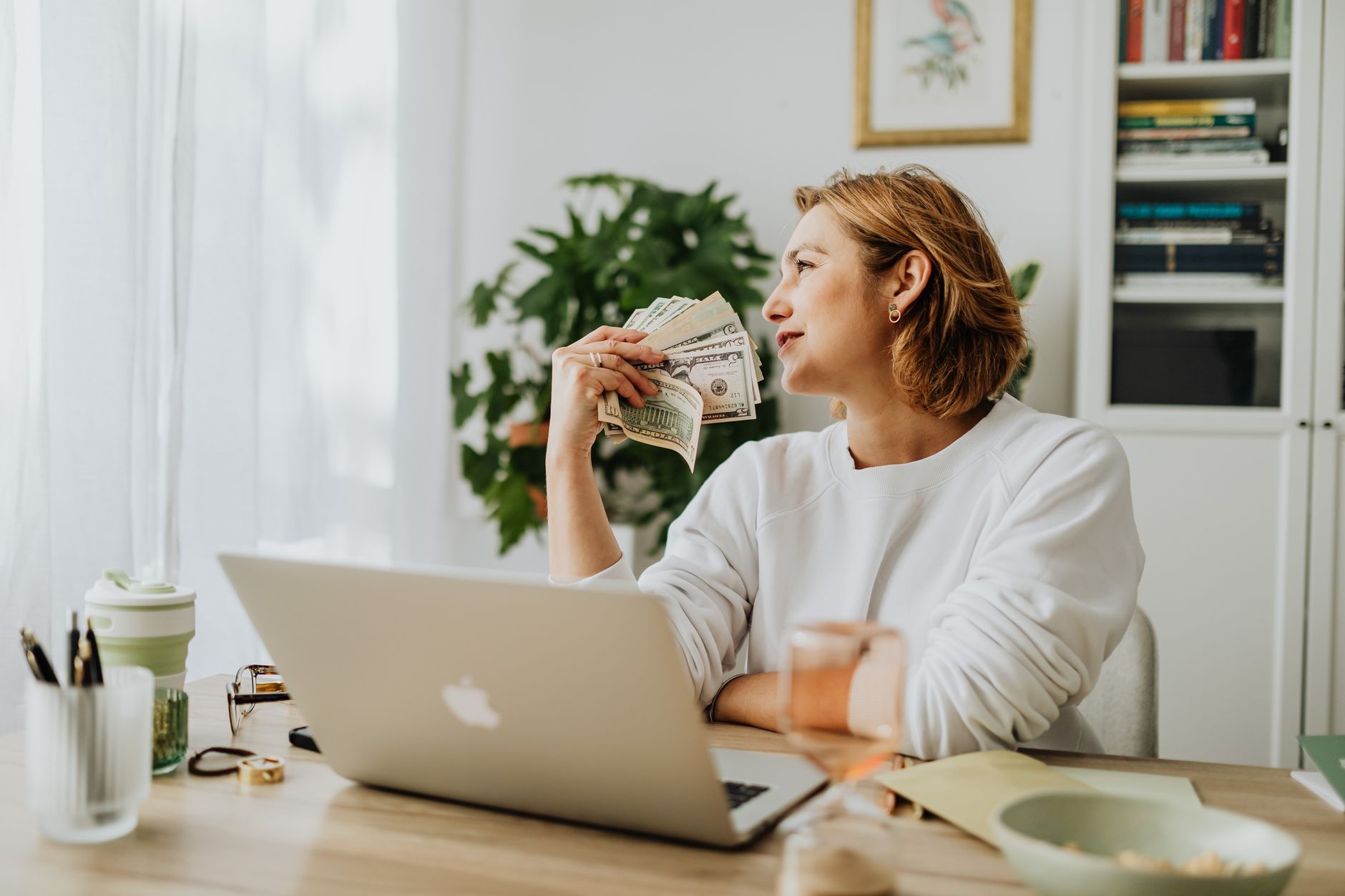 A woman at a desk with a laptop holding cash and looking thoughtfully away for a side hustle concept.