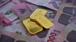Three gold bars resting on a scattered pile of pink Indonesian rupiah banknotes.
