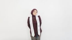 A female graduate in a dark purple hijab and white sash poses happily against a white wall.