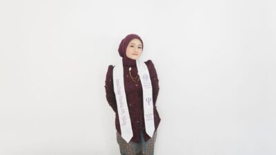 A female graduate in a dark purple hijab and white sash poses happily against a white wall.