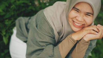 A smiling young woman in a hijab rests her chin in her hands outdoors, exemplifying the benefits of smiling.