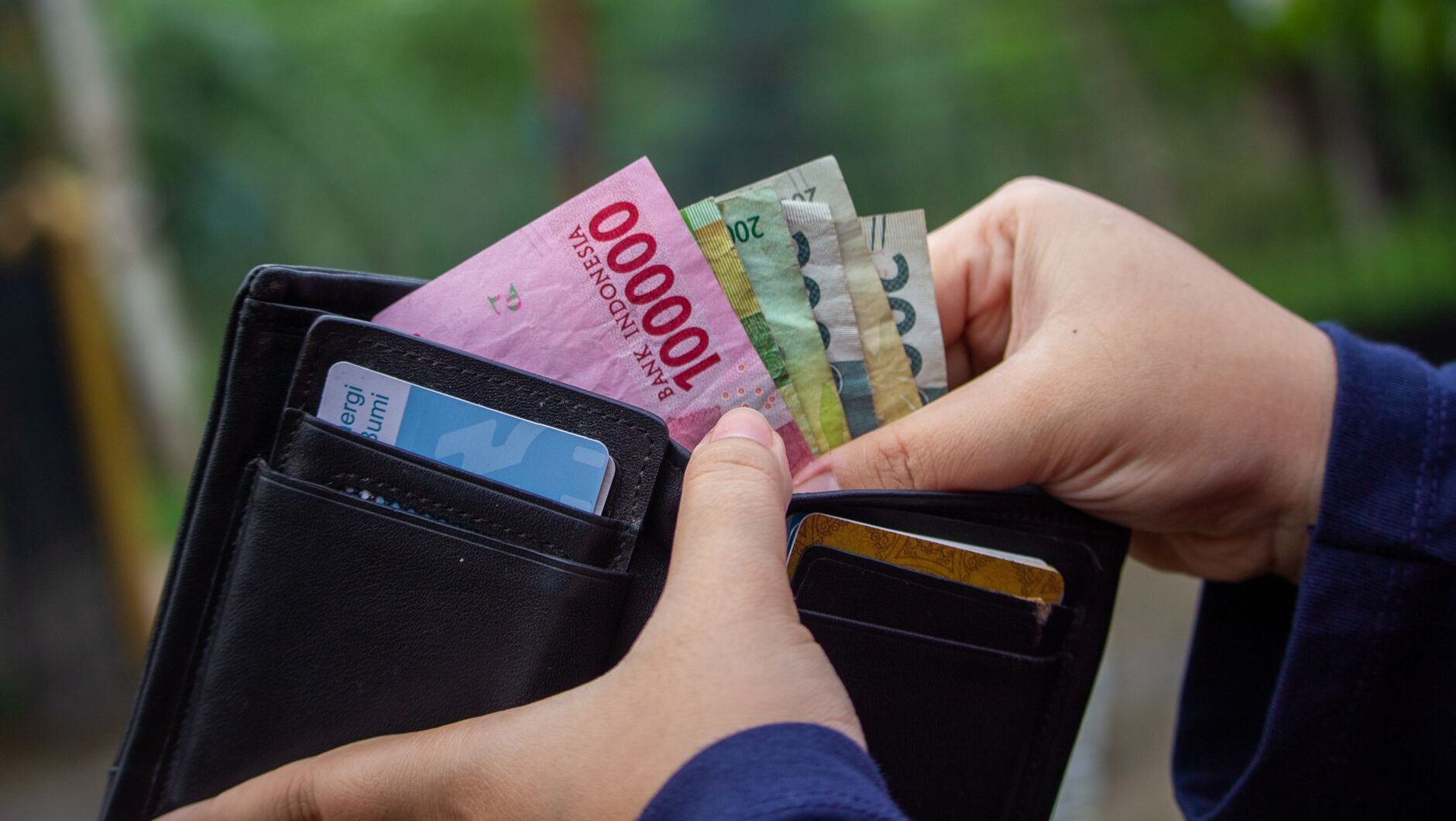Seseorang mengeluarkan uang Rupiah dari dompet kulit hitam berisi kartu kredit.