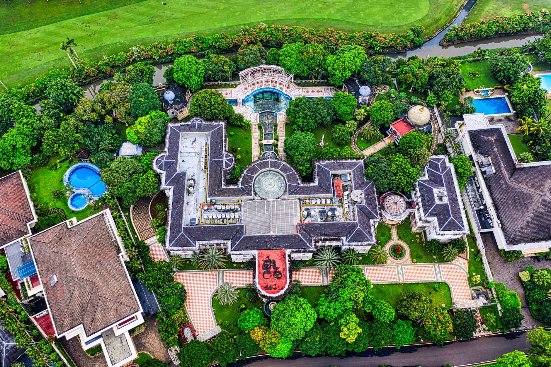 Overhead aerial view of a sprawling luxury mansion with a swimming pool and lush landscaped gardens, representing Biaya Pensi
