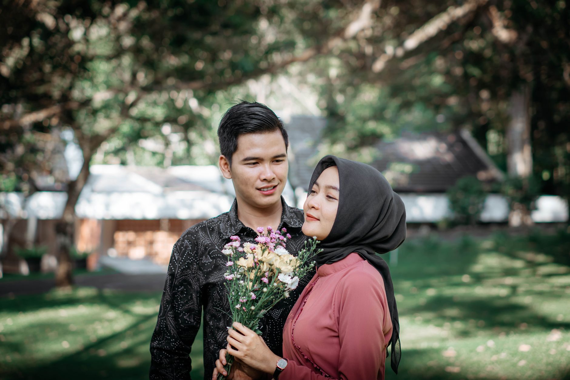 A smiling man stands beside a woman in a hijab holding a bouquet of flowers in a lush green garden.