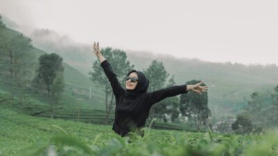 Seorang wanita berhijab hitam dan berkacamata hitam merentangkan tangan di kebun teh yang asri untuk menumbuhkan rasa percaya