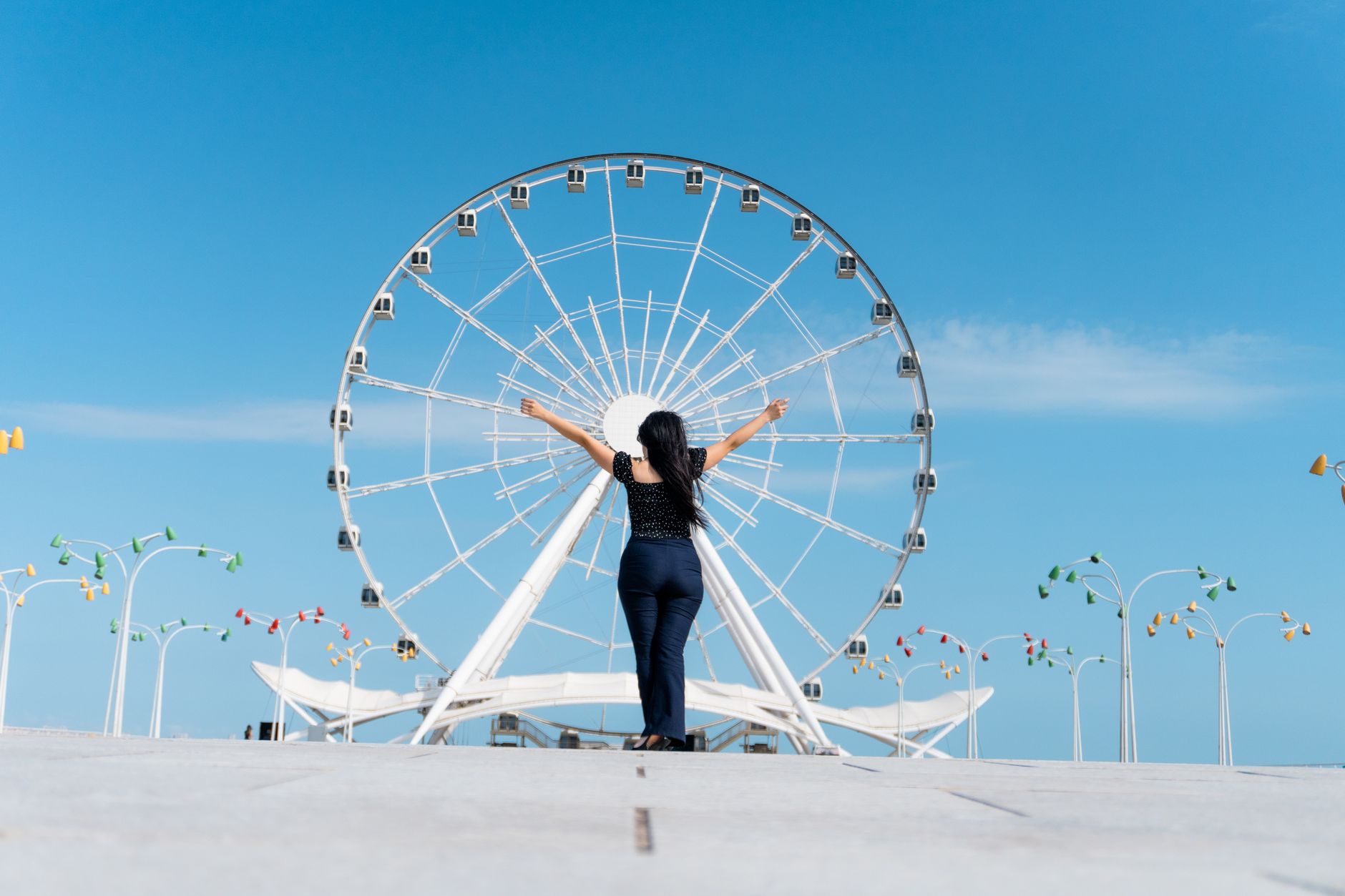 A woman with arms outstretched facing a giant Ferris wheel under a clear blue sky, radiating Energi Positif Attraction.