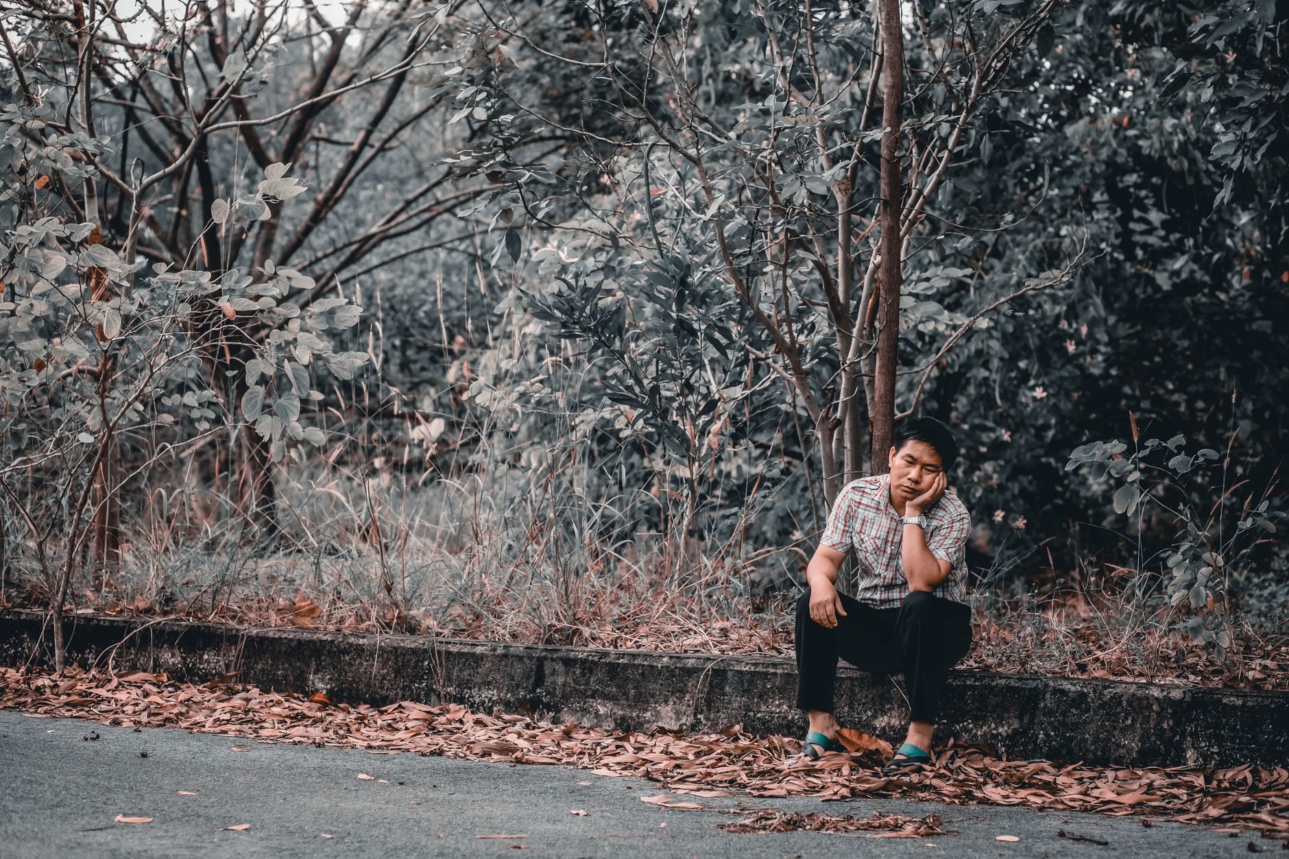 Seated man looking thoughtful on a roadside curb near a lush forest, representing Mengatasi Kesepian.