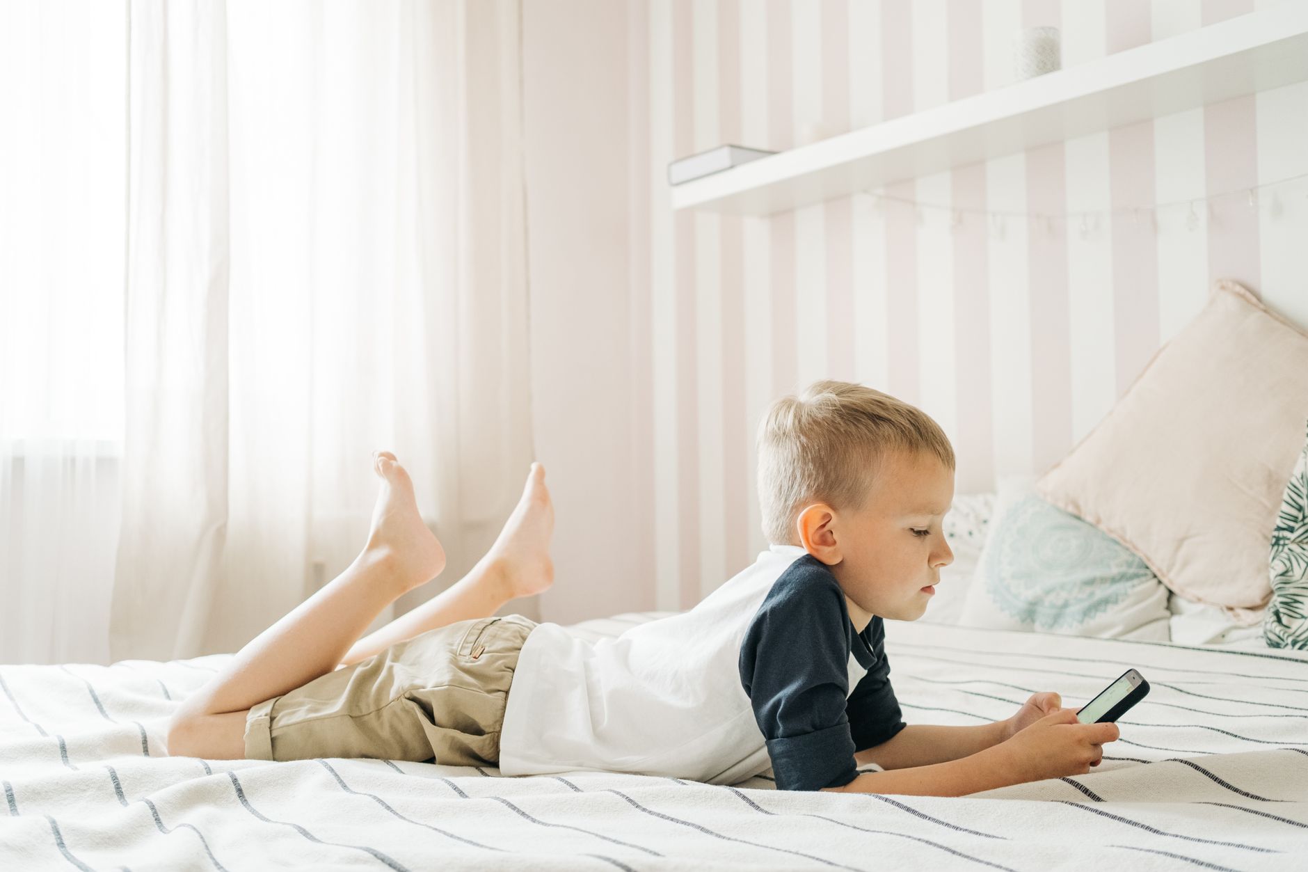 Seorang anak laki-laki berbaring di tempat tidur sambil asyik bermain game di smartphone, menggambarkan dampak screen time an