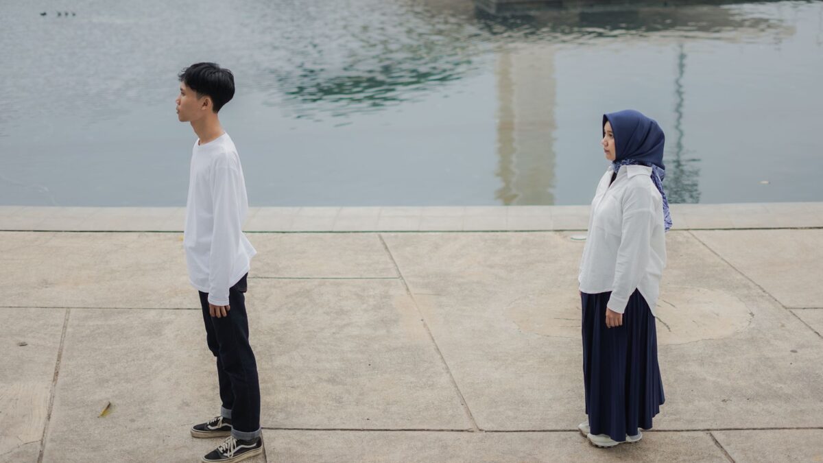 A young man and woman stand back-to-back on a pier, representing a pasangan tidak serius in a relationship.