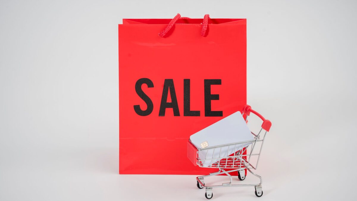 A red sale shopping bag behind a mini shopping cart holding credit cards.