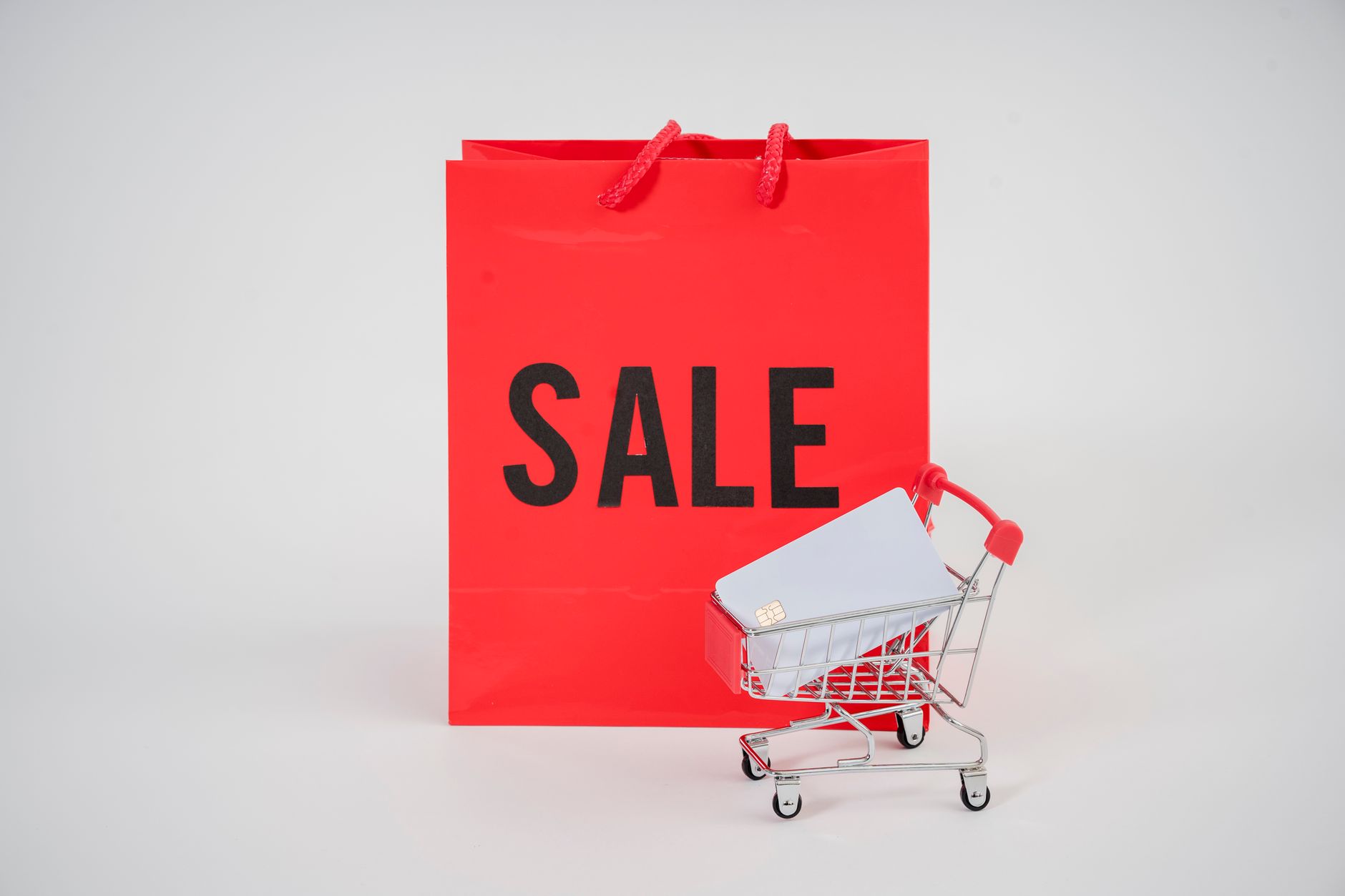 A red sale shopping bag behind a mini shopping cart holding credit cards.
