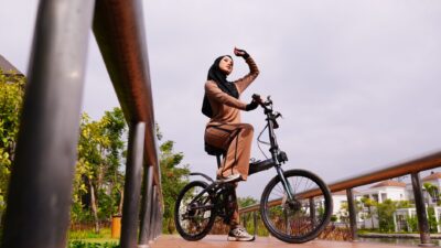 Seorang wanita berhijab mengenakan pakaian cokelat berpose di atas sepeda di jembatan kayu saat siang hari.