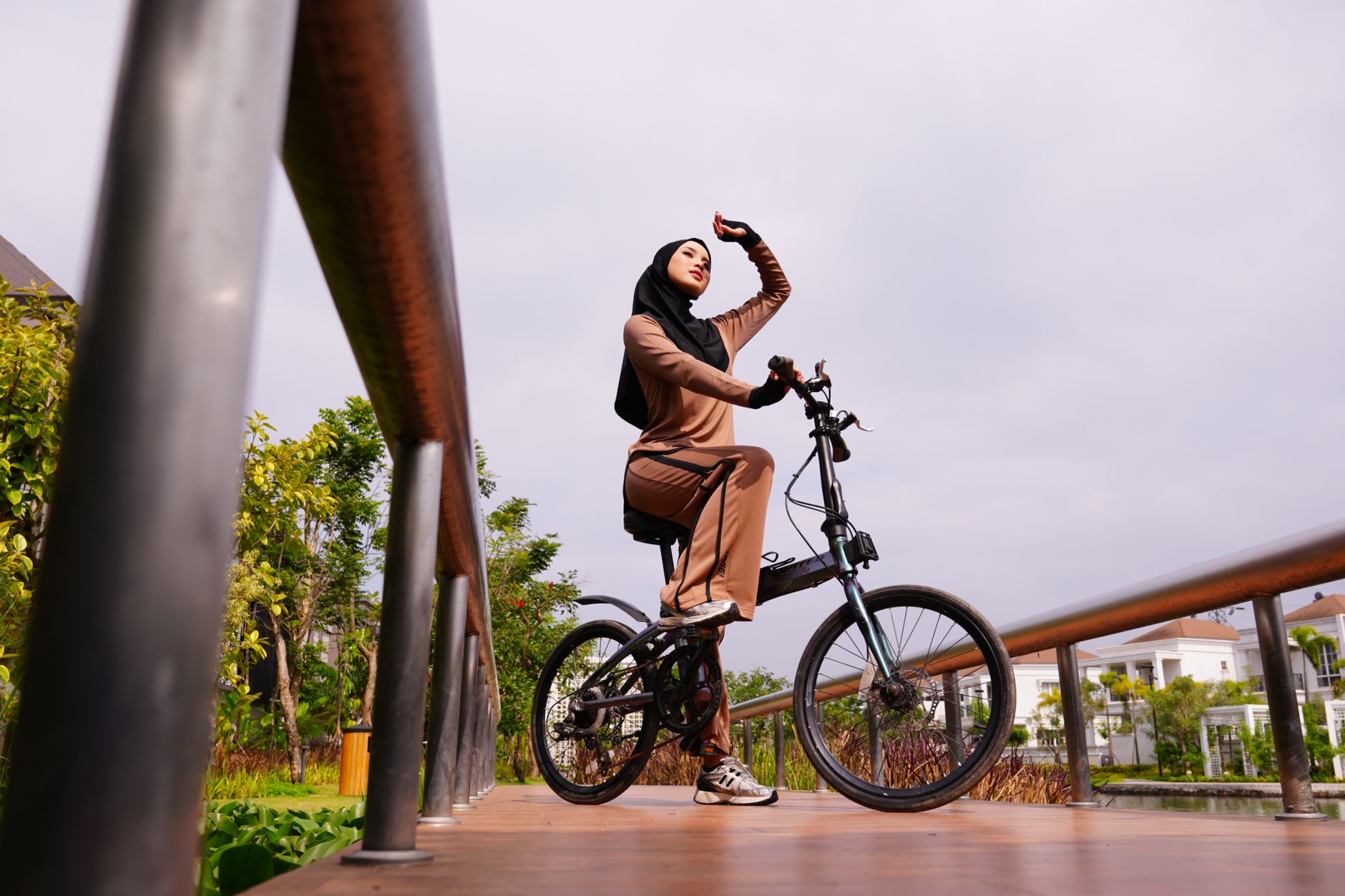 Seorang wanita berhijab mengenakan pakaian cokelat berpose di atas sepeda di jembatan kayu saat siang hari.