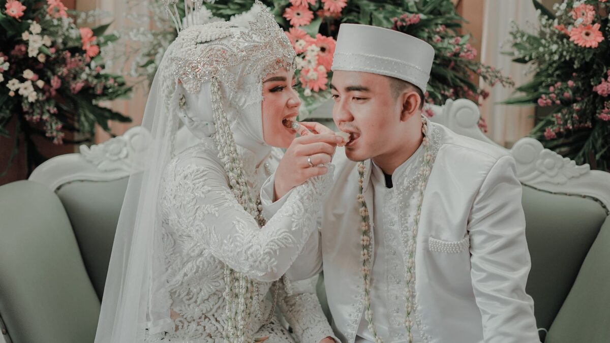Indonesian bride and groom in white traditional attire smiling while feeding each other sweets at their wedding.