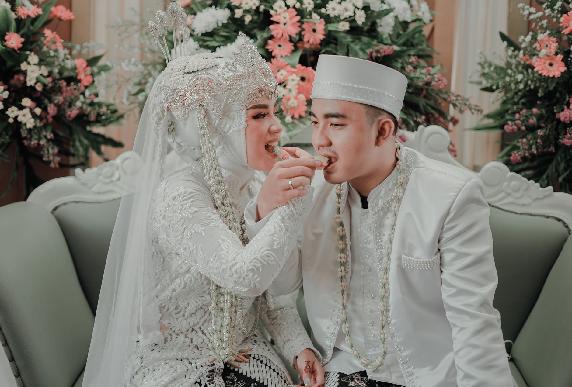 Indonesian bride and groom in white traditional attire smiling while feeding each other sweets at their wedding.