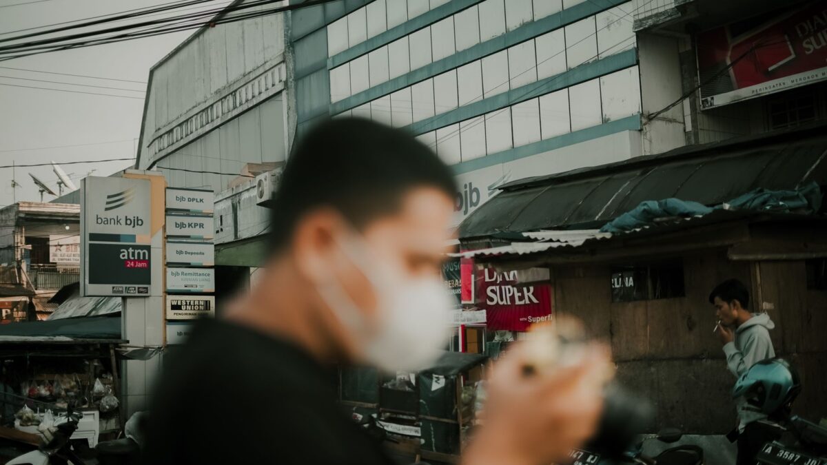 A person in a face mask walks down a busy city street past a bank bjb ATM sign.